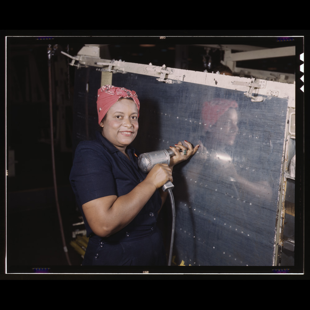 African American aircraft assembler in blue coveralls and red bandana working on a Vengence dive bomber at the Vultee-Nashville plant, 1943.]